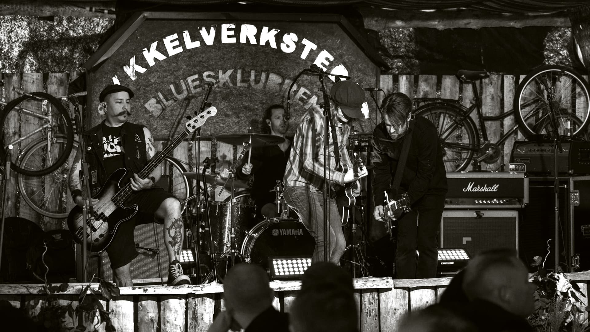 Band på scenen i Sykkelverkstedet, med sykler hengende på veggen bak. Svart-hvitt.